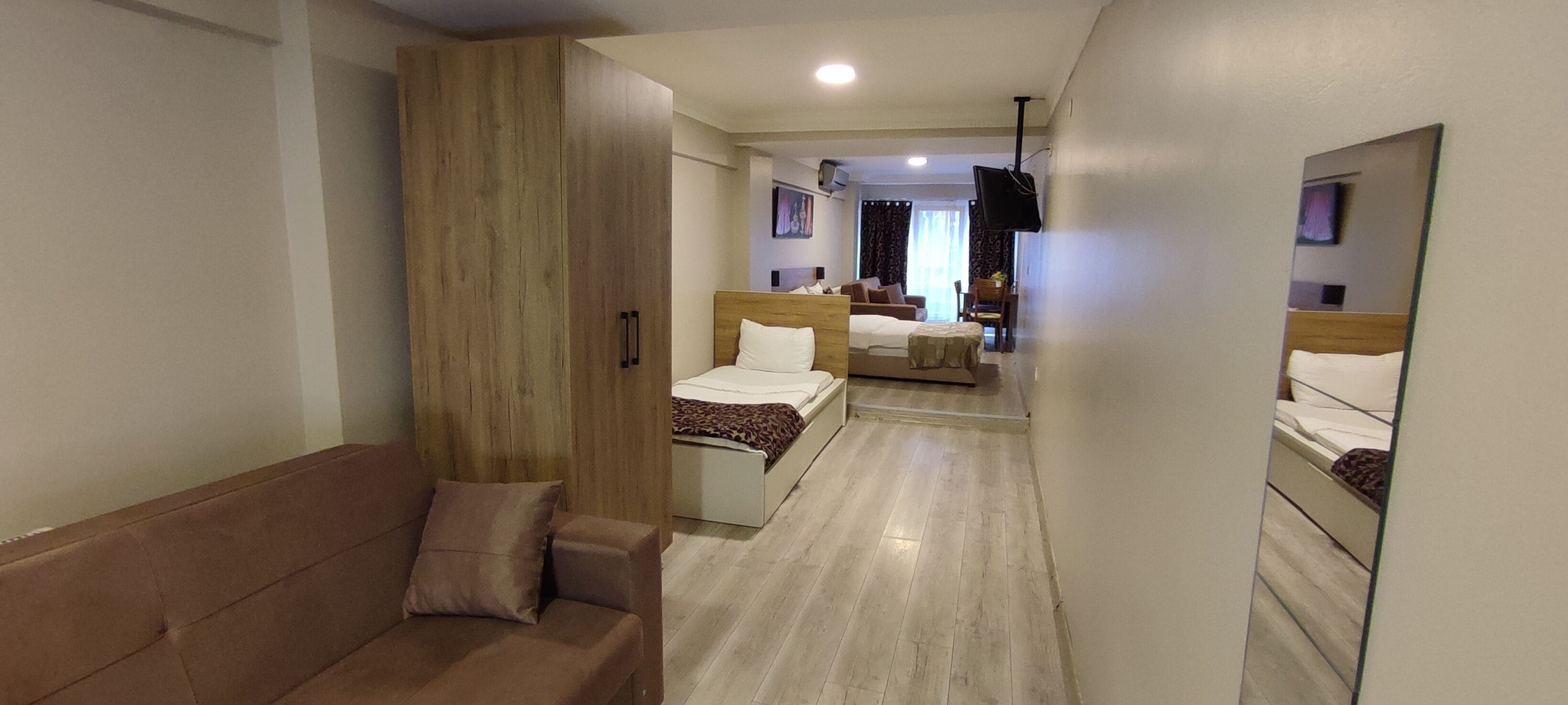 Una camera d'albergo con un armadio in legno, un divano marrone, due letti, una televisione montata a parete e un corridoio che porta a un'altra stanza.
