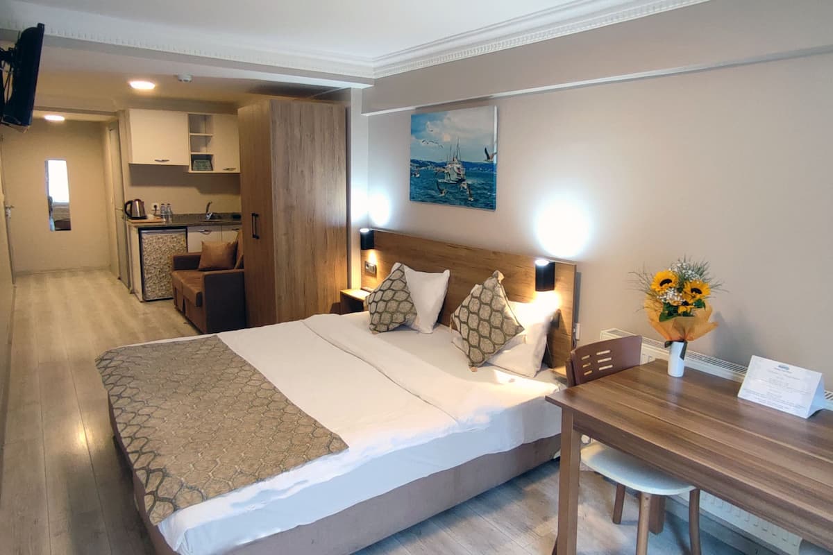 A hotel room with a bed, a wooden desk, a chair, a small table, a vase with flowers, a painting on the wall, and a kitchenette in the background.