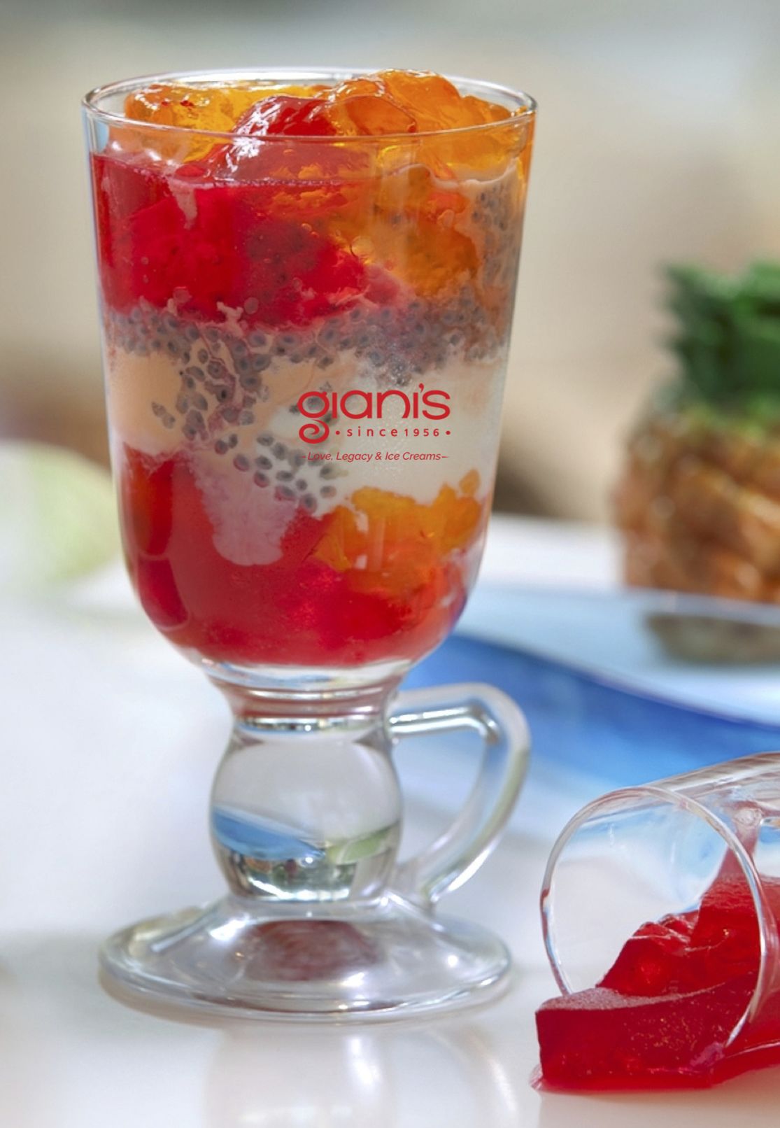 Un dessert servi dans un verre transparent muni d’une anse, composé de couches de gelée rouge, de crème blanche et de petites garnitures rondes, le tout portant la marque « giannis depuis 1956 ».