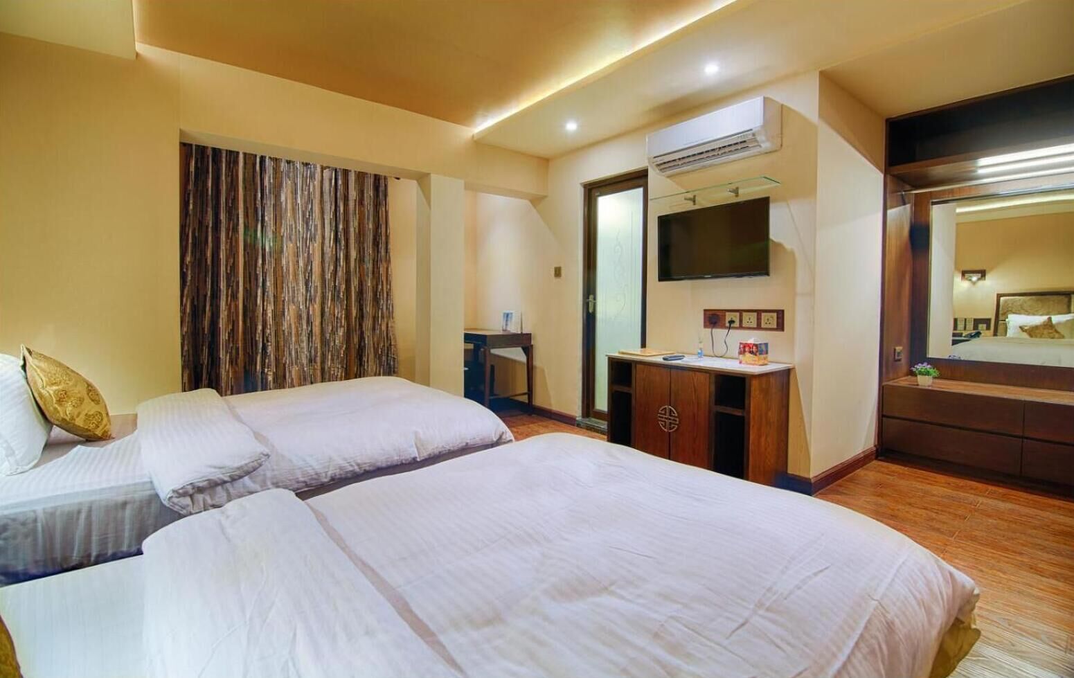 A hotel room with two beds, a desk, a TV, and an air conditioning unit.