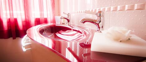 A modern bathroom with a red sink, chrome faucets, and a white rose on a napkin.