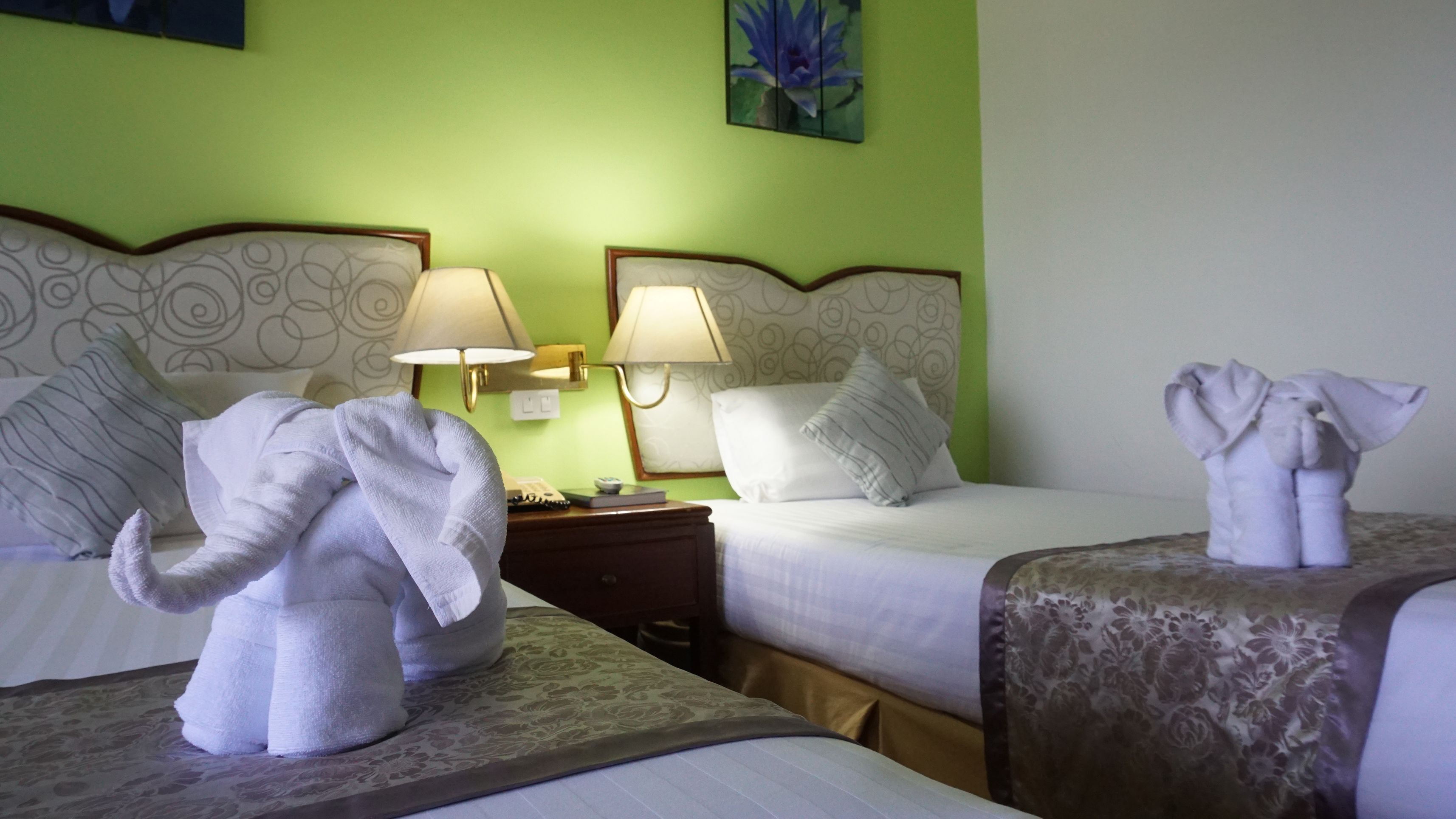 A hotel room with two beds, each with a headboard, a nightstand with a lamp, and decorative towels shaped like elephants.