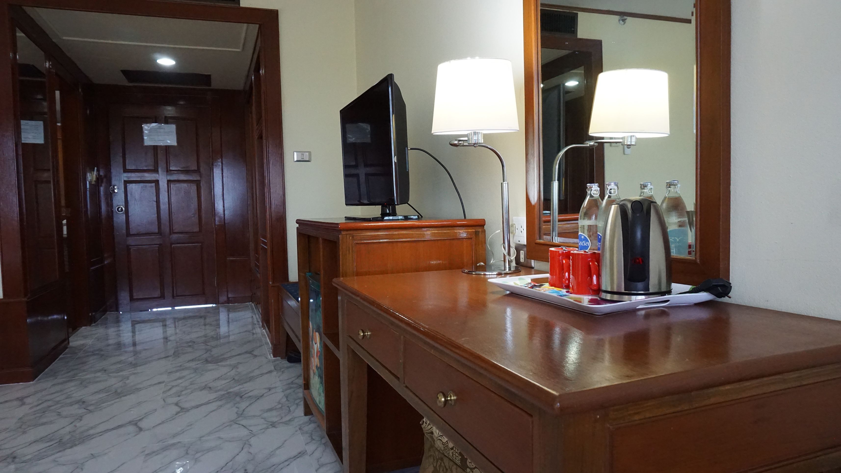 A hotel room with a wooden dresser, a television, two bedside lamps, a mirror, and a tray with cups and a kettle.
