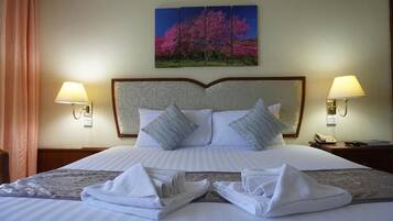 Habitación de hotel con una cama grande, dos lámparas de noche, un cuadro de árboles rosados y toallas dobladas con cuidado sobre la cama.