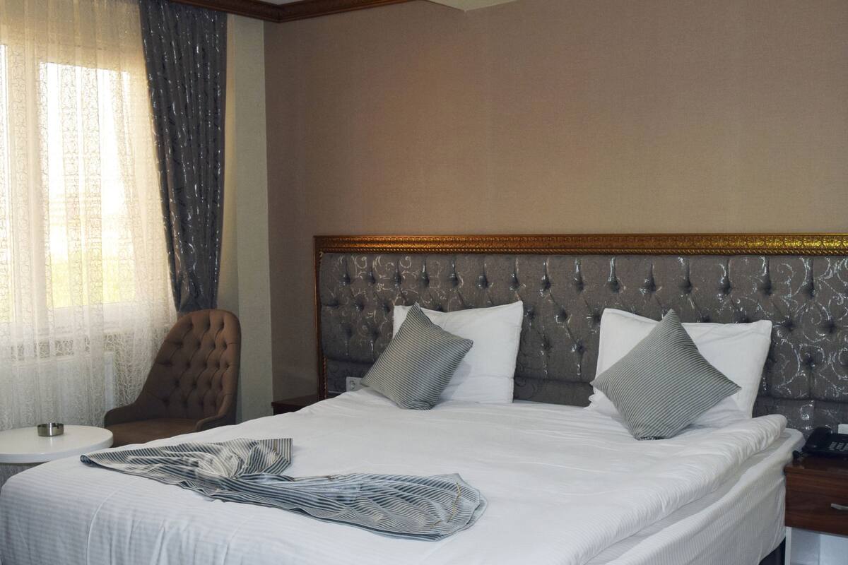 A hotel room with a large bed, a chair, a bedside table, and a window with curtains.