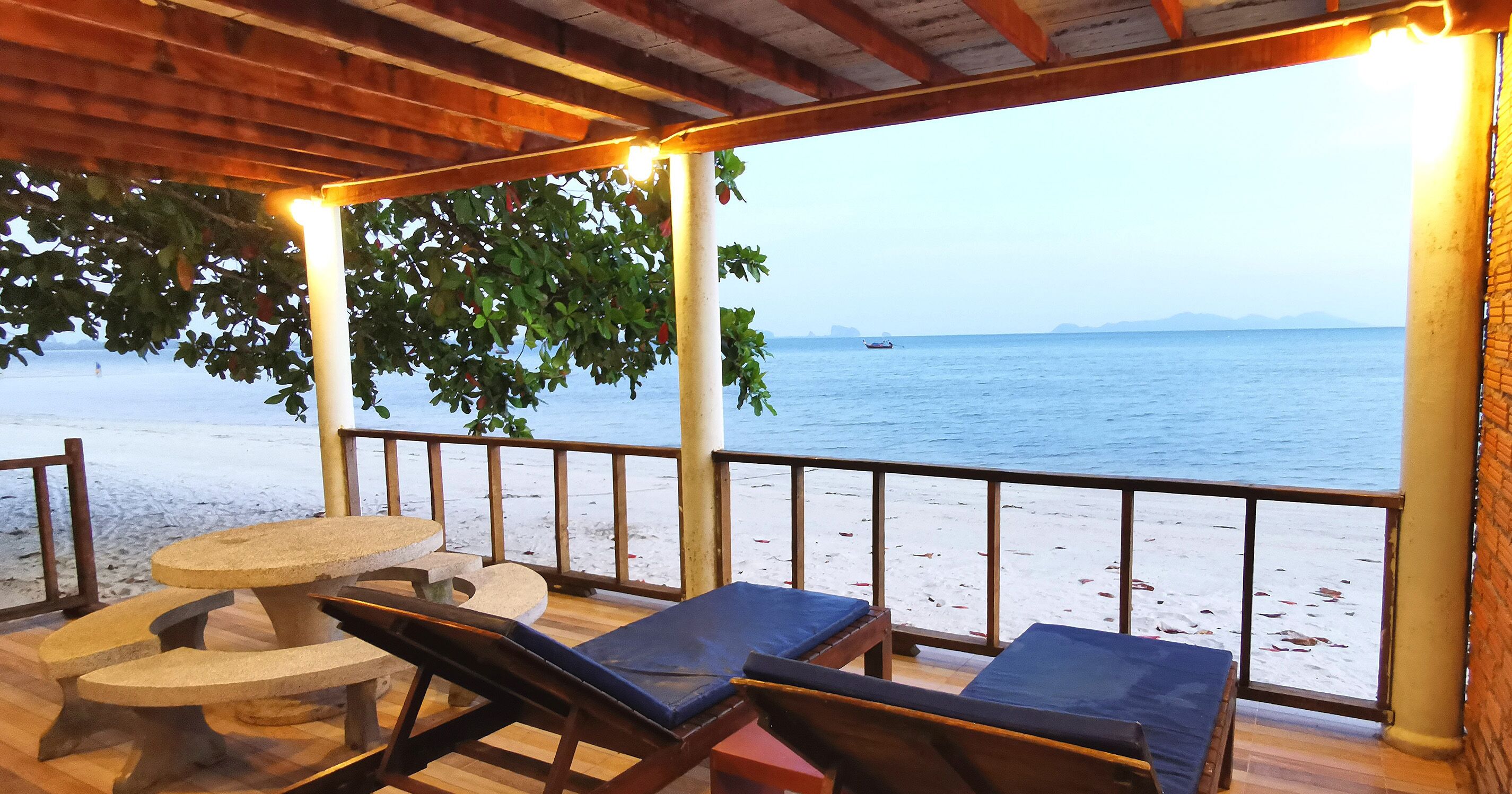 A covered outdoor seating area with a table and lounge chairs, overlooking a beach with a boat in the distance.