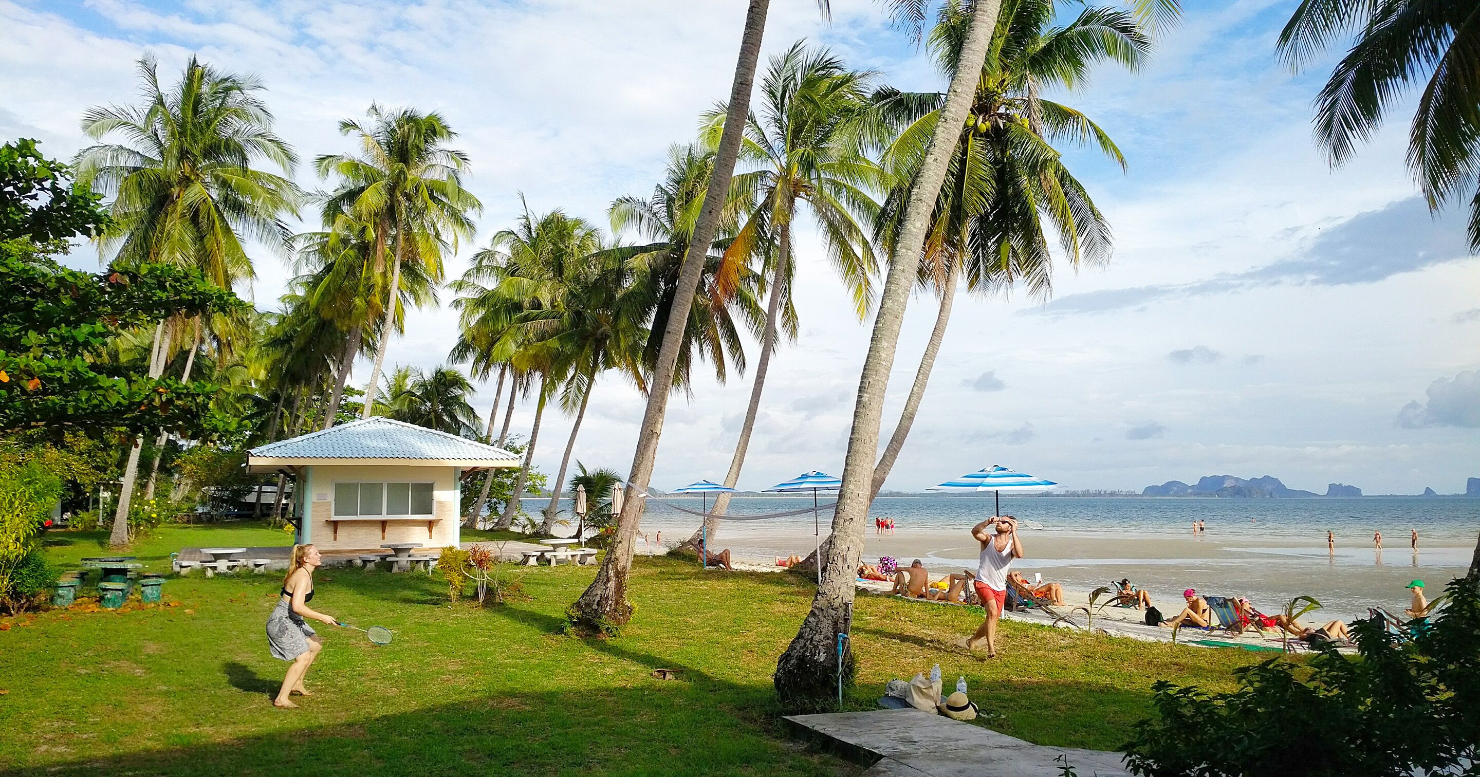A beachside scene with palm trees, a small building, and people relaxing on the sand.