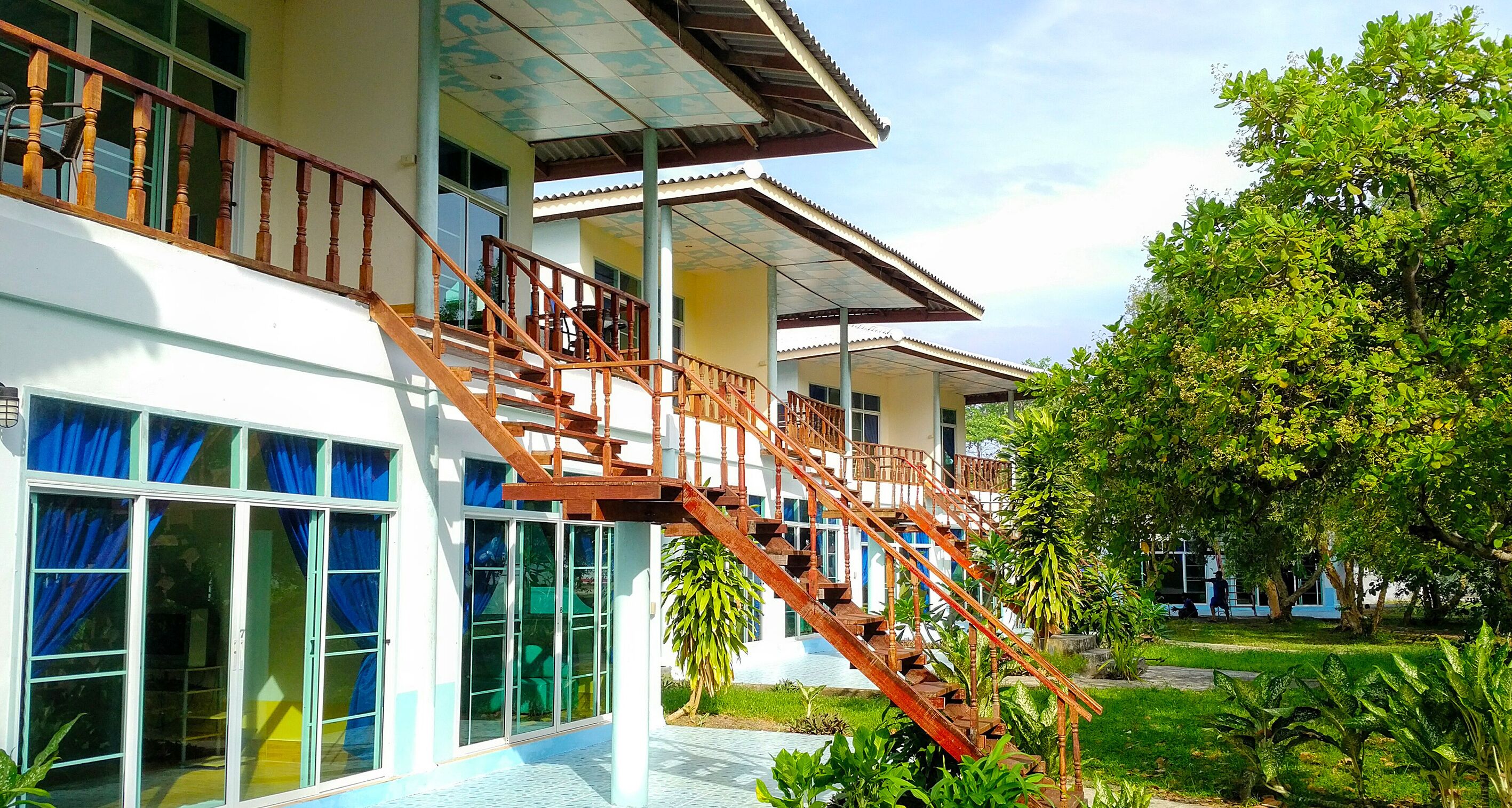 A building with multiple floors, a staircase with wooden railings, large windows with blue curtains, and a garden area with various plants.
