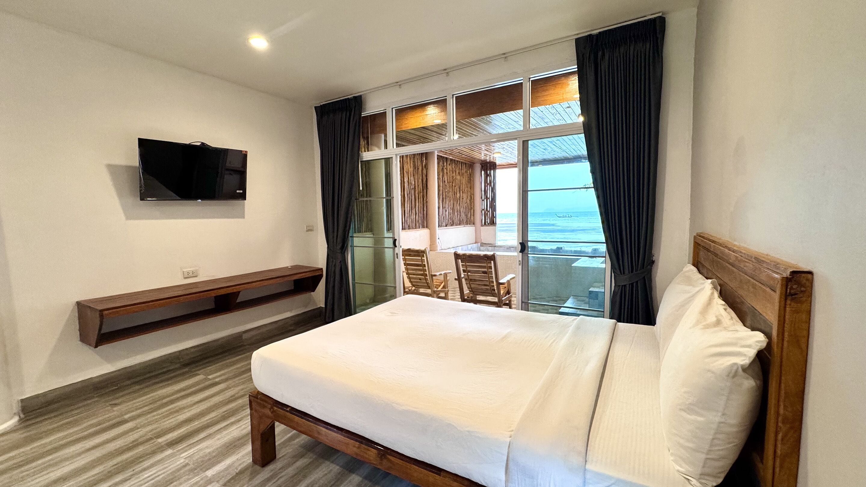 A hotel room with a bed, a TV mounted on the wall, a bench, and a view of the ocean through large windows.