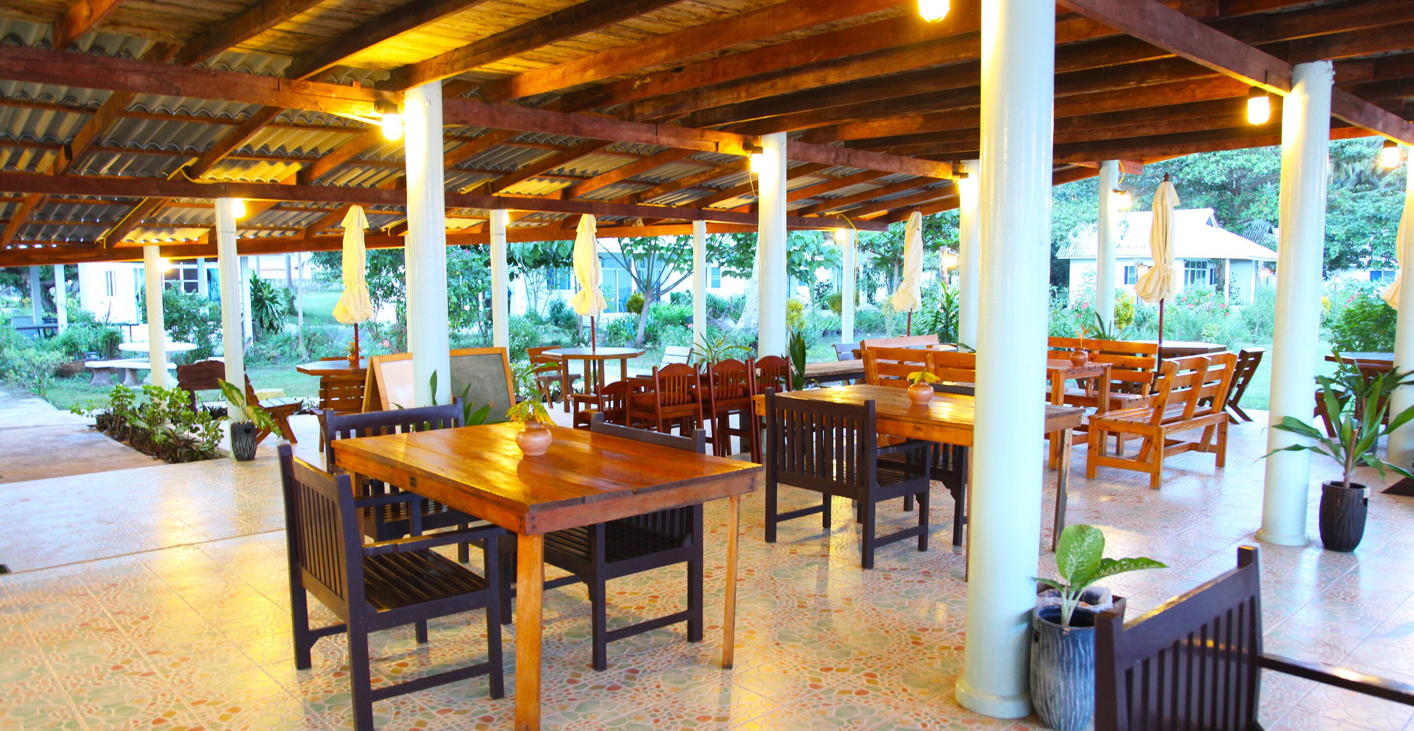 An outdoor covered patio area with wooden tables and chairs, potted plants, and a view of greenery.