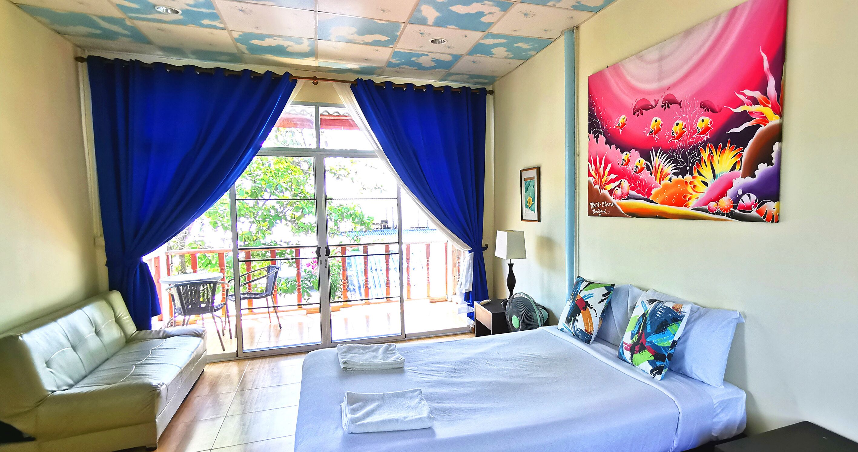 A hotel room with a bed, a sofa, a fan, a window with blue curtains, and a colorful painting on the wall.