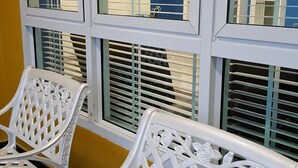 Zwei weiße Stühle mit aufwendigen Gittermustern stehen vor einem Fenster mit horizontalen Rollläden.