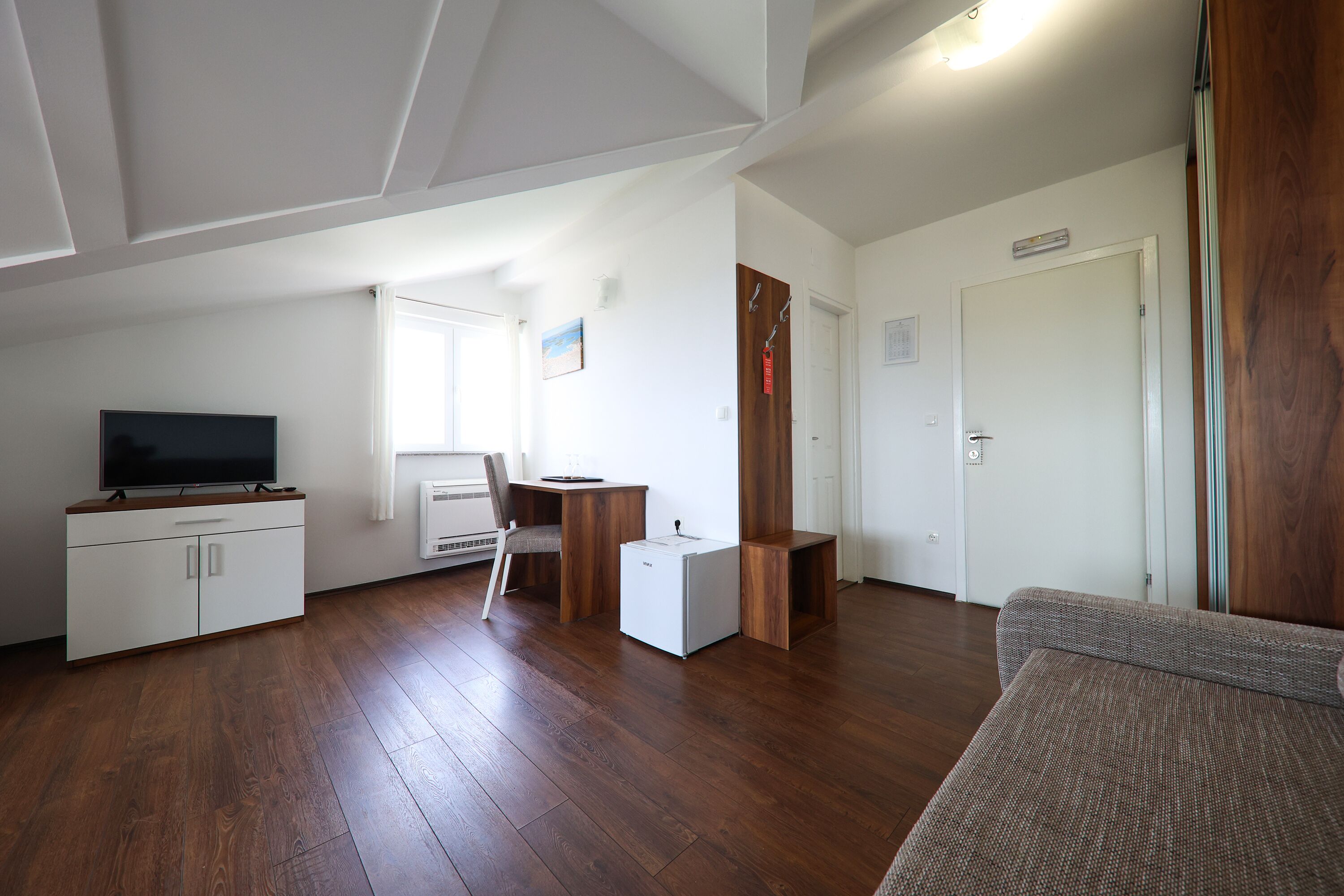 A modern living room with a flat-screen TV, wooden flooring, a white cabinet, a small white refrigerator, and a grey sofa.