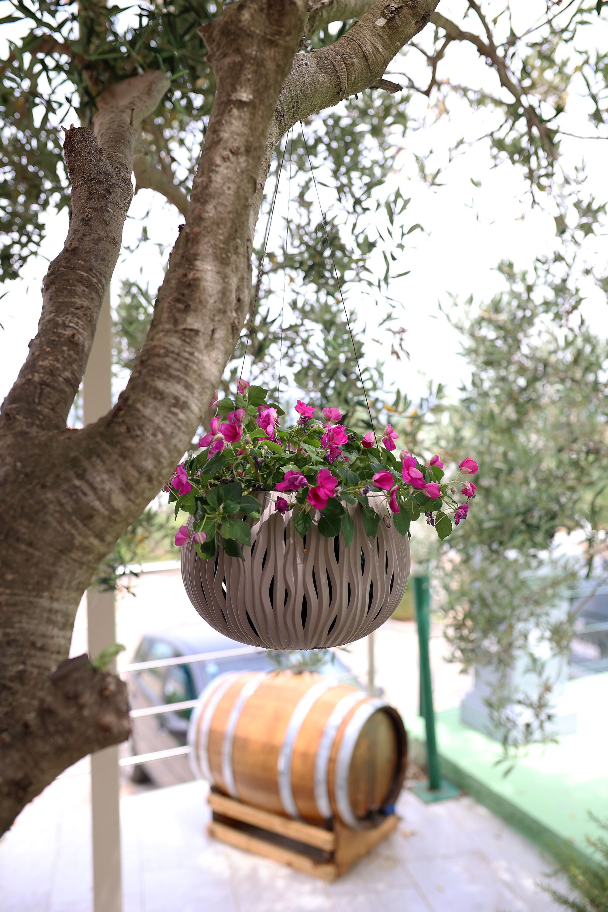 A hanging flower pot with pink flowers, a wooden barrel, and a tree trunk.