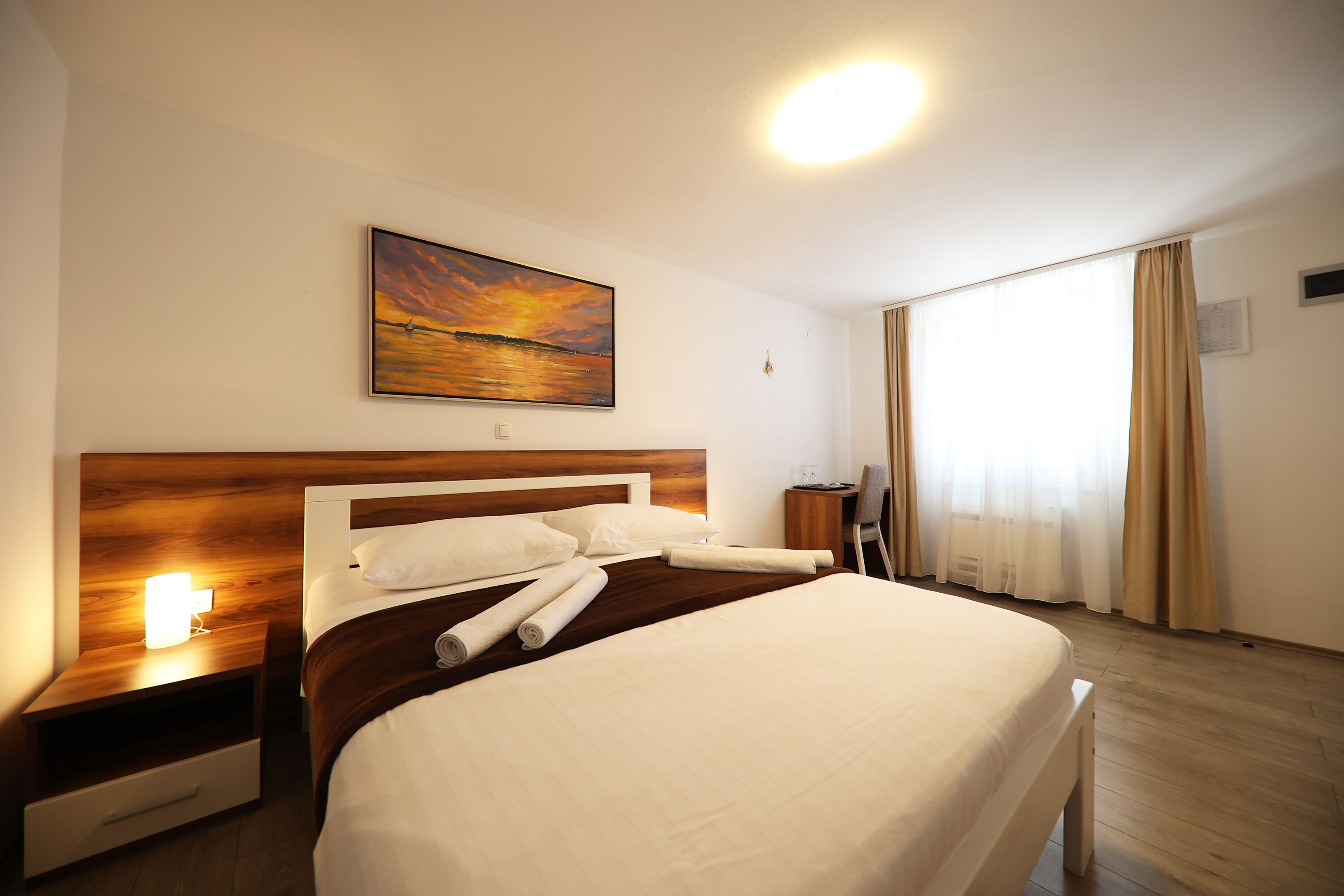 A hotel room with a large bed, a nightstand with a lamp, a painting of a sunset, and a window with curtains.