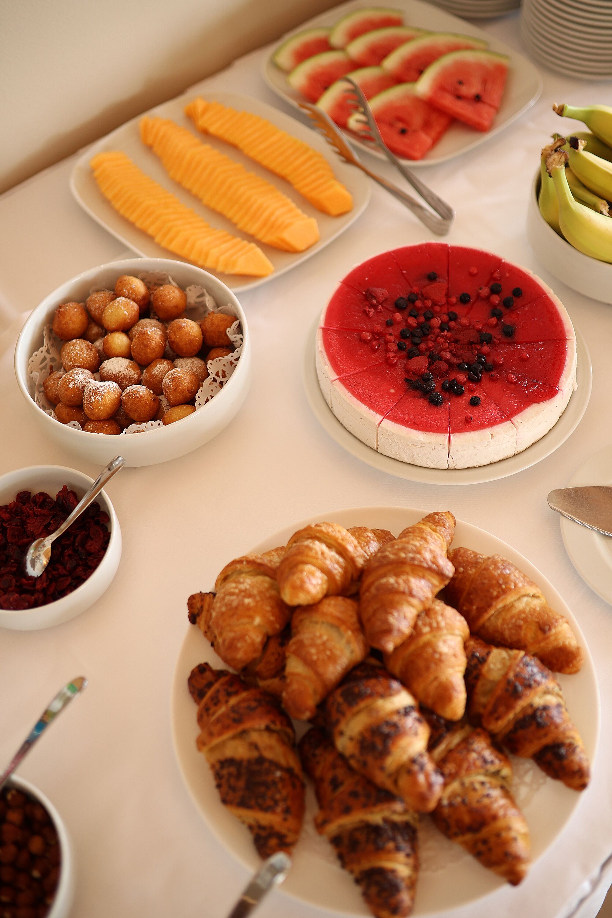 A table with various food items including watermelon slices, bananas, croissants, and a cake.