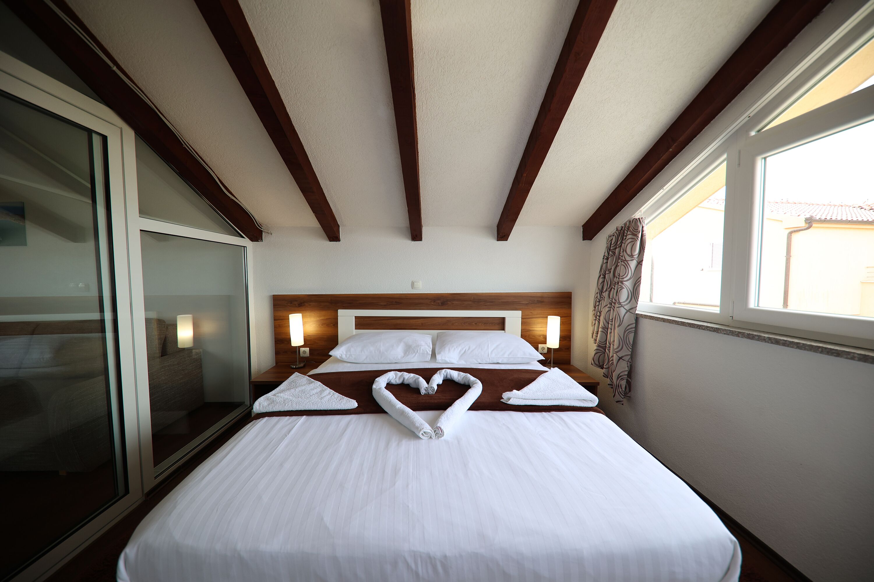 Habitación de hotel con una cama grande, vigas de madera en el techo, un ventanal amplio y una disposición de toallas en forma de corazón sobre la cama.