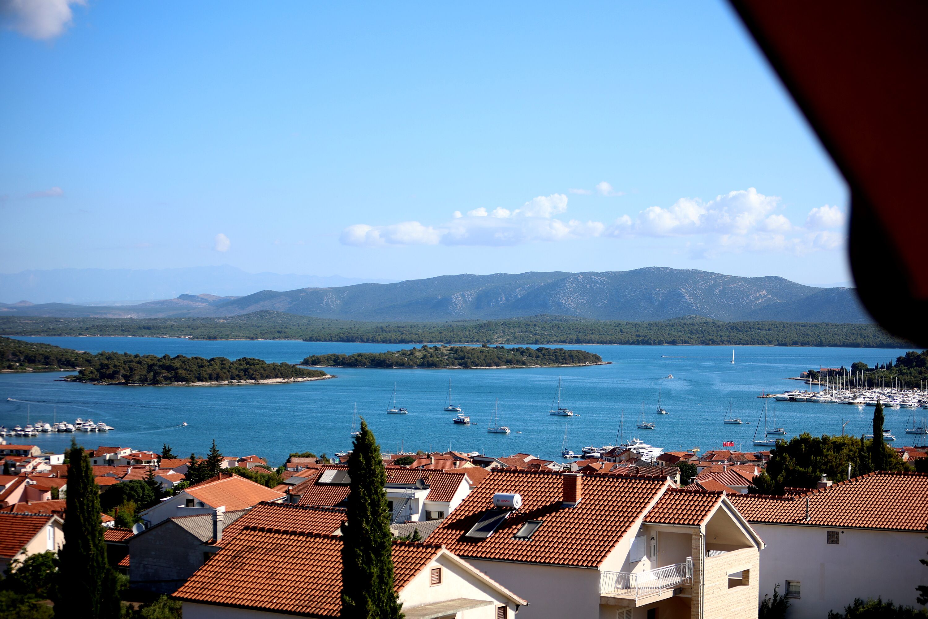 A coastal town with terracotta-roofed buildings, a marina with boats, and a clear view of the sea and distant mountains.