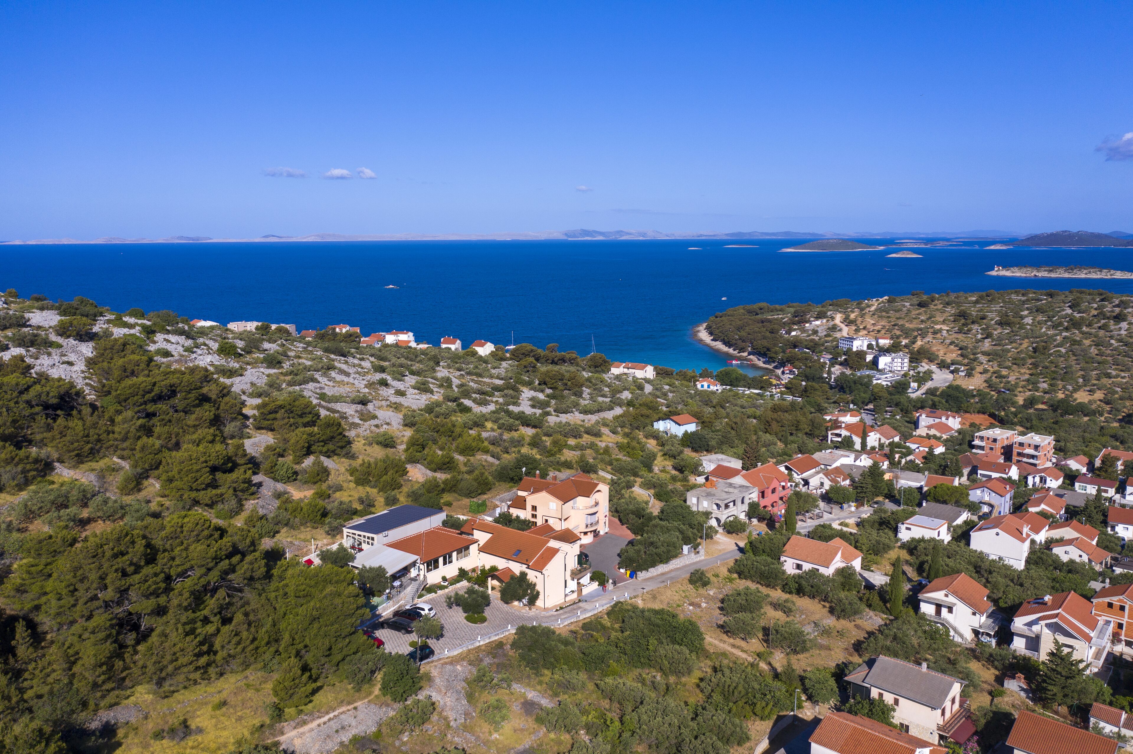 Luftaufnahme einer Küstenwohngegend mit Häusern, tiefblauem Meer und einer hügeligen Landschaft.