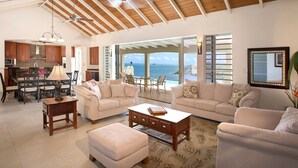 Un vaste espace de vie avec une grande fenêtre donnant sur la mer, un coin repas avec une table et des chaises, et une cuisine dotée de meubles en bois et d’un ventilateur de plafond.