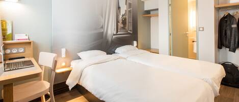 A hotel room with a bed, a desk with a laptop, a chair, and a coat rack with jackets.