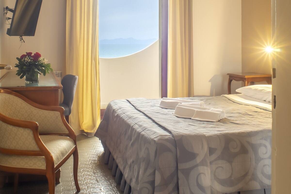 Habitación de hotel con cama, escritorio con monitor de computadora, una silla, un florero con flores y vista al mar.