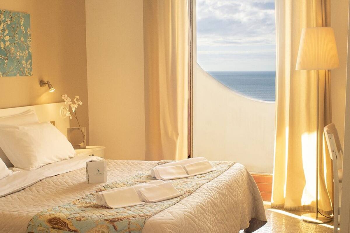 Habitación de hotel con cama, mesita de noche, lámpara y vista al mar.
