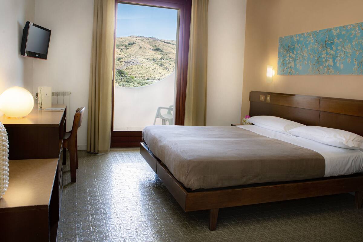 Habitación de hotel con cama, escritorio, silla y vista a un paisaje montañoso a través de una ventana.