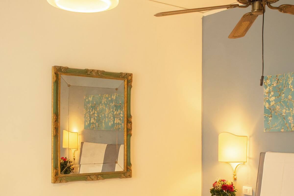 Habitación de hotel con cama, ventilador de techo, espejo, mesita de noche con lámpara y un cuadro floral en la pared.