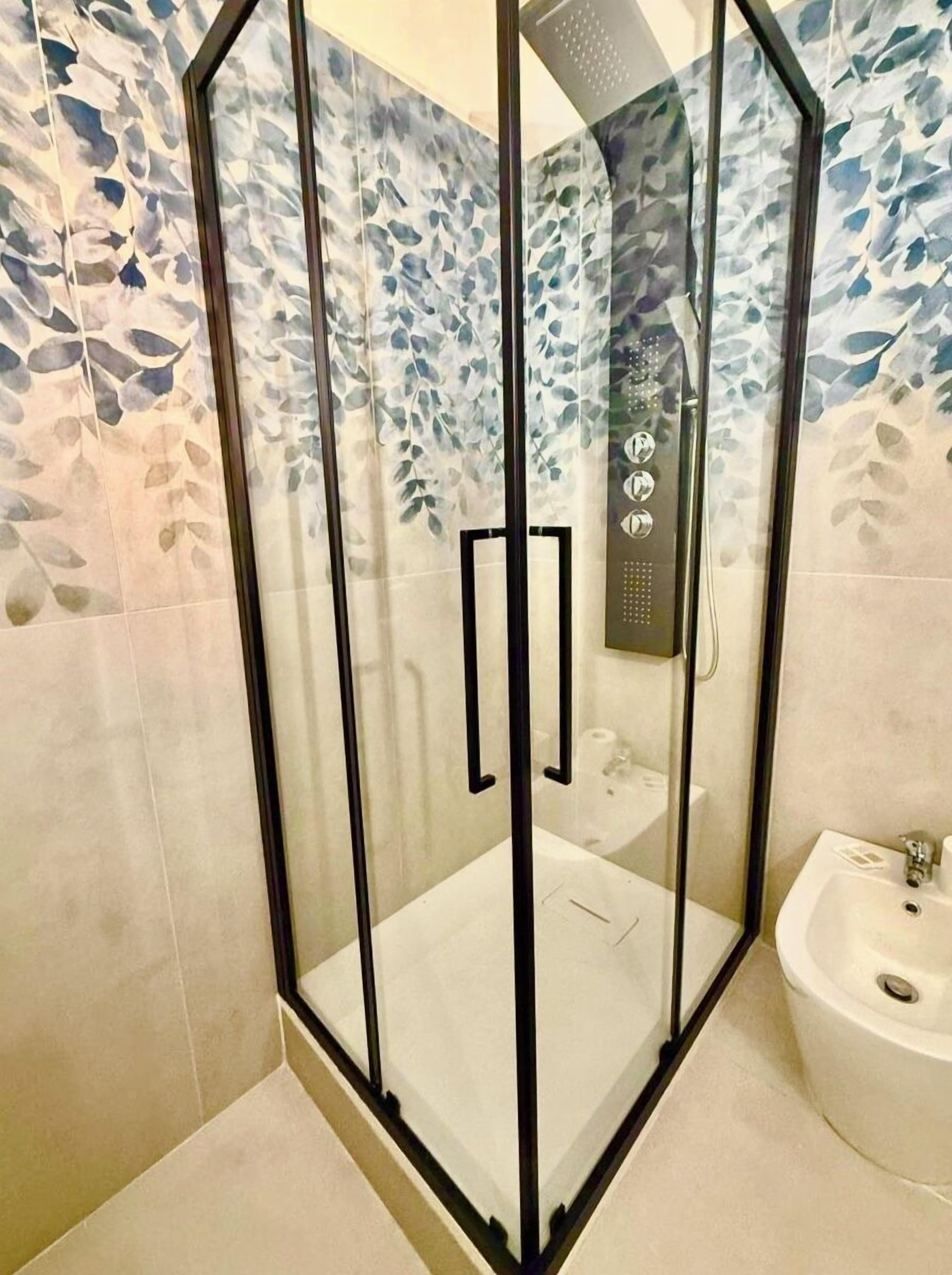 A glass-enclosed shower with a control panel, a white sink, and a tiled wall with a leaf pattern.