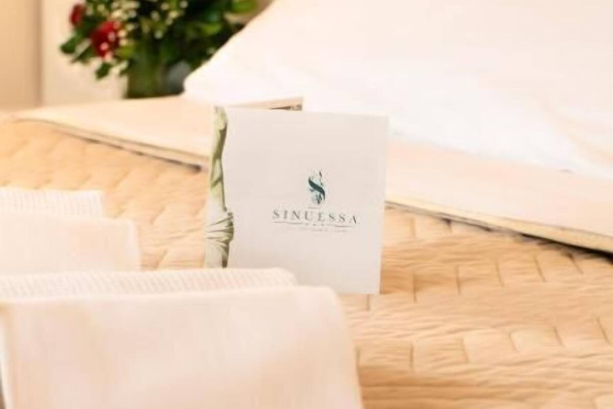 Una cama bien hecha con cabecera blanca, una tarjeta con el nombre 'Sinuessa' y un ramo de flores rojas sobre la mesita de noche.