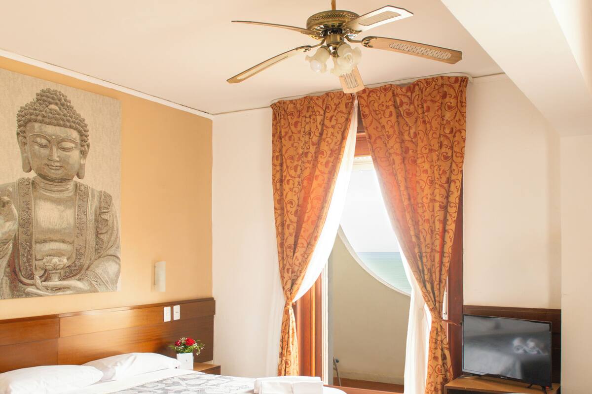 Habitación de hotel con dos camas, ventilador de techo, estatua de Buda, televisor y vistas al océano a través de cortinas.