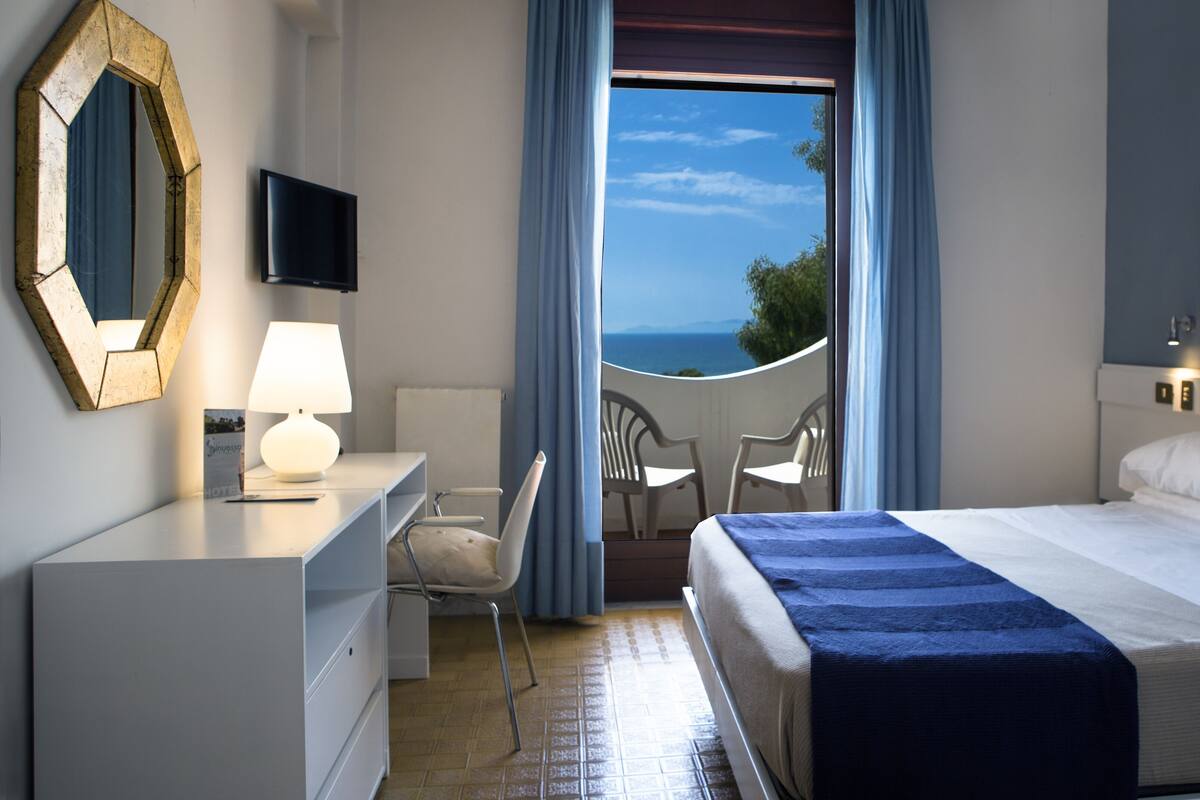 Una habitación de hotel con cama, escritorio con lámpara, silla, balcón con vista al mar y un espejo grande en la pared.