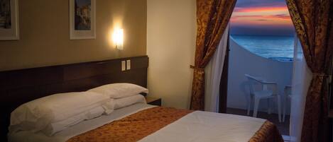 A hotel room with a bed, a nightstand, a wall-mounted light, a view of the ocean through curtains, and a chair by the window.
