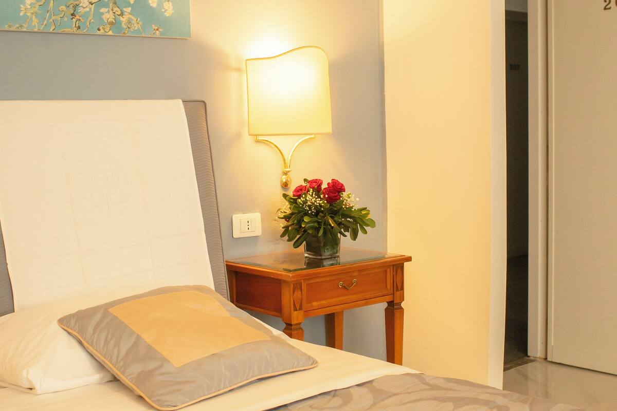 Una cama bien hecha con un edredón estampado, una mesita de noche con una lámpara y un jarrón de flores, y una pared con un cuadro floral.