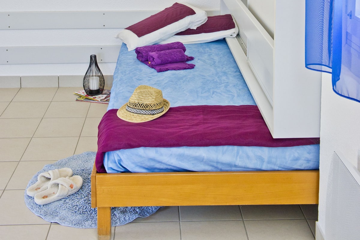 A single bed with a wooden frame, a blue mattress, and a purple blanket. There is a small vase on the floor beside the bed.