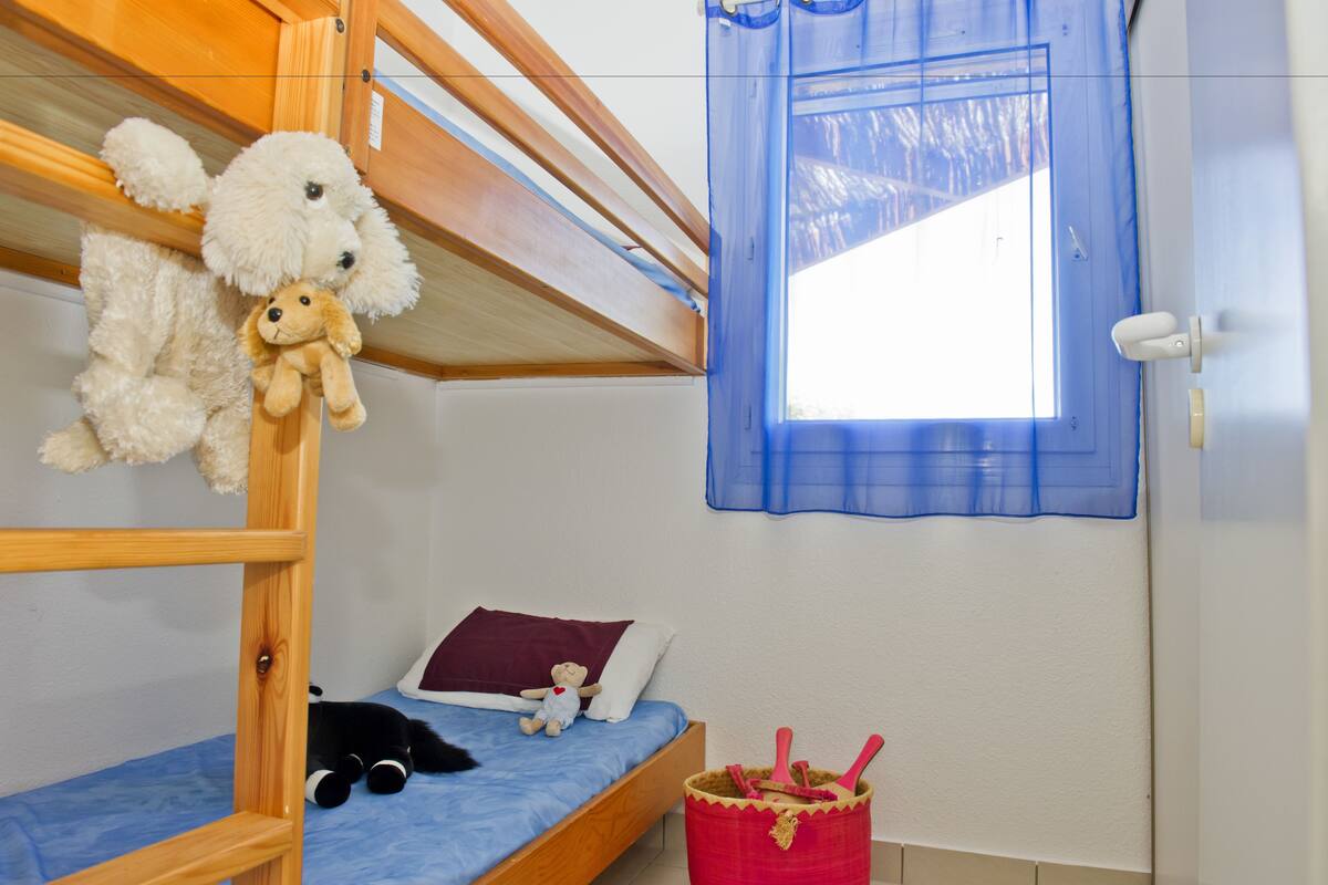 A bunk bed with a wooden ladder, a small bedside table, a red trash bin, and a window with blue curtains.