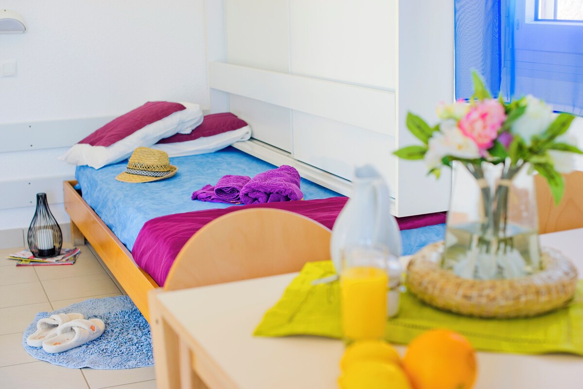 A neatly arranged bedroom with a bed, a bedside table with a vase of flowers, a bottle of orange juice, and a pair of slippers on a blue mat.