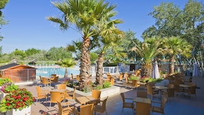 An outdoor patio area with tables and chairs, palm trees, and a pool in the background. - Azureva Le Grau du Roi (Le Grau-Du-Roi)