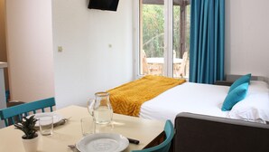 A hotel room with a bed, a TV mounted on the wall, a dining table with chairs, a small plant, and a window with curtains. - Azureva Le Grau du Roi (Le Grau-Du-Roi)