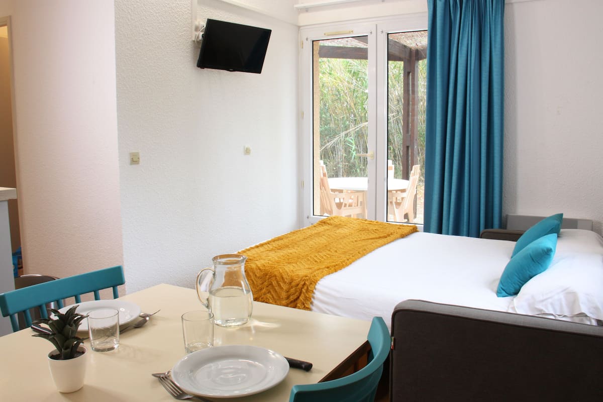 A hotel room with a bed, a TV mounted on the wall, a dining table with chairs, a small plant, and a window with curtains.