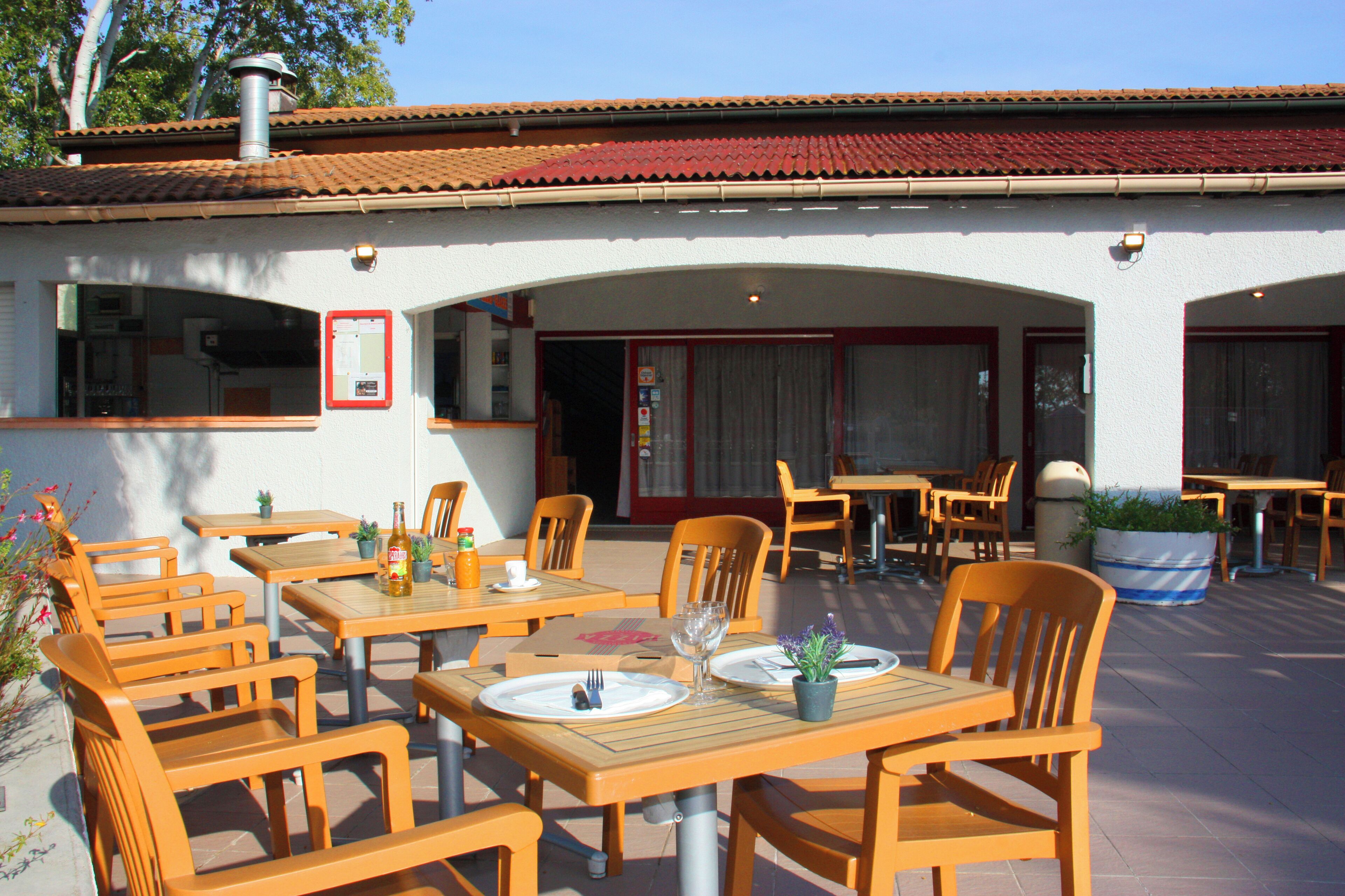 An outdoor patio with wooden tables and chairs, a potted plant, and a trash can.
