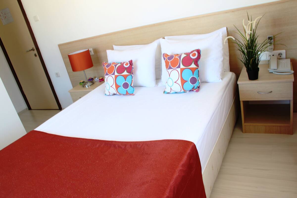 A hotel room with a large bed, two pillows with a floral pattern, a bedside table with a lamp, a phone, and a small plant.