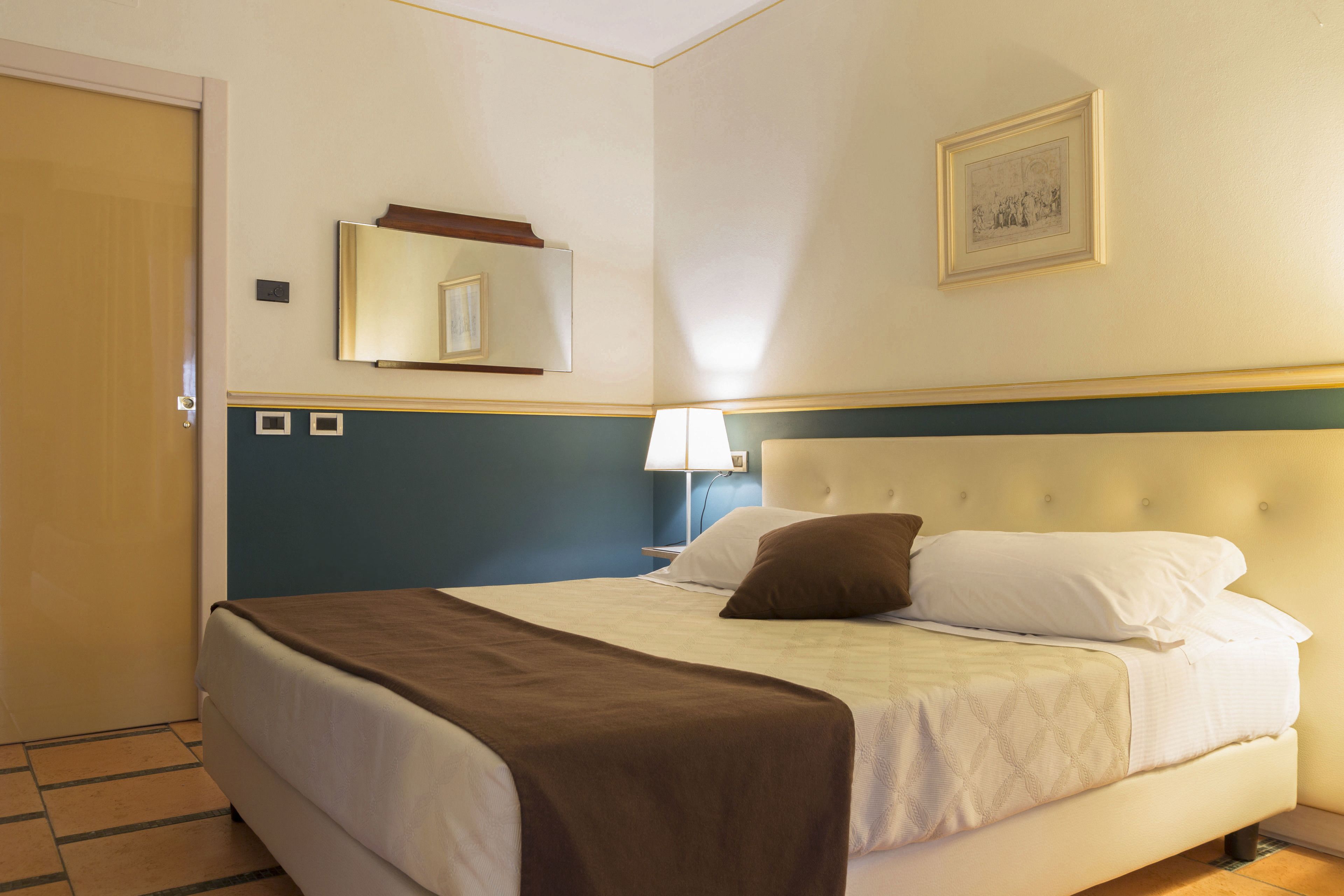 Ein Hotelzimmer mit einem großen Bett, einem Nachttisch mit Lampe, einem Spiegel und einem gerahmten Bild an der Wand.