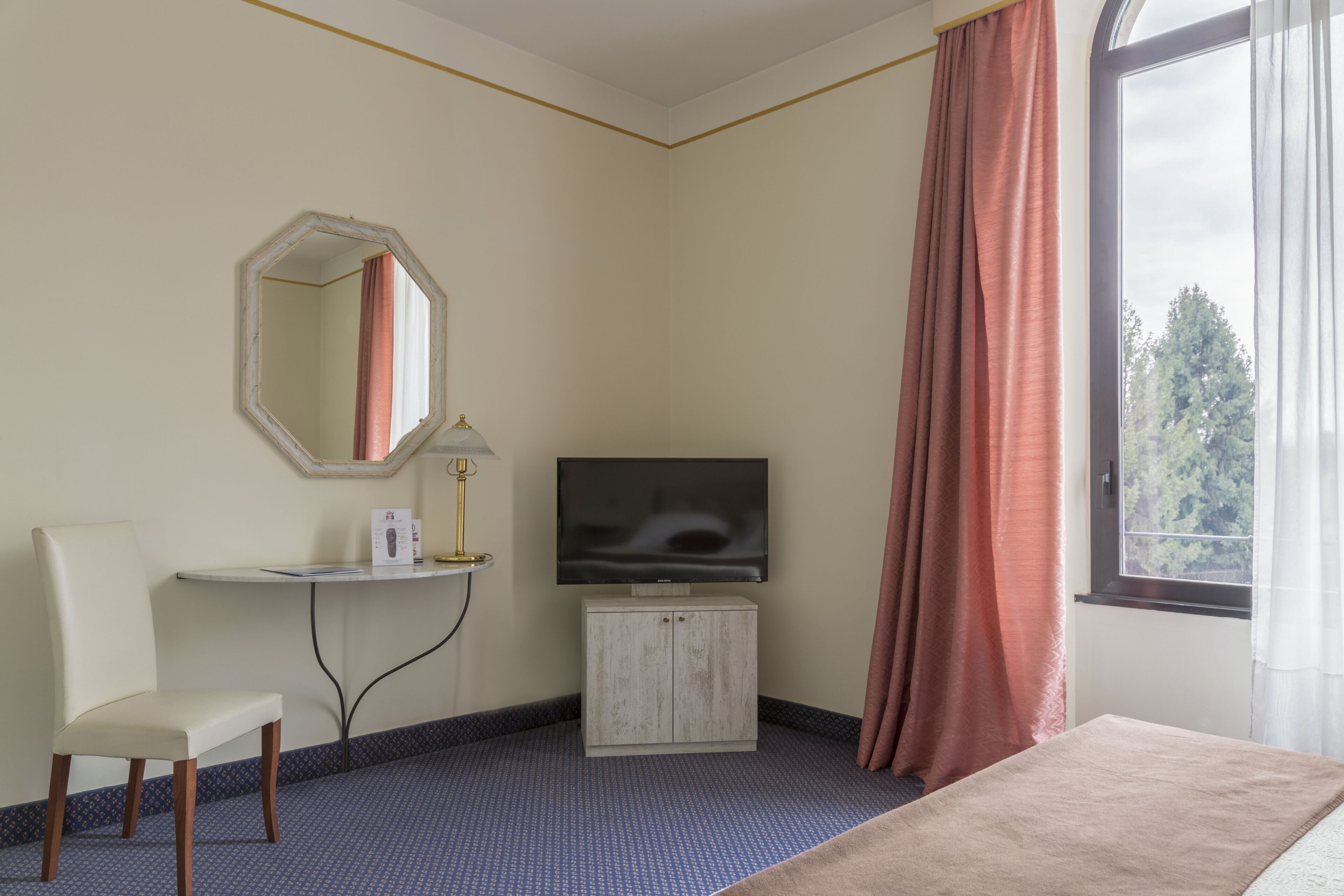 Habitación de hotel con cama, televisor sobre un mueble bajo, una silla, un escritorio y una ventana con cortinas.