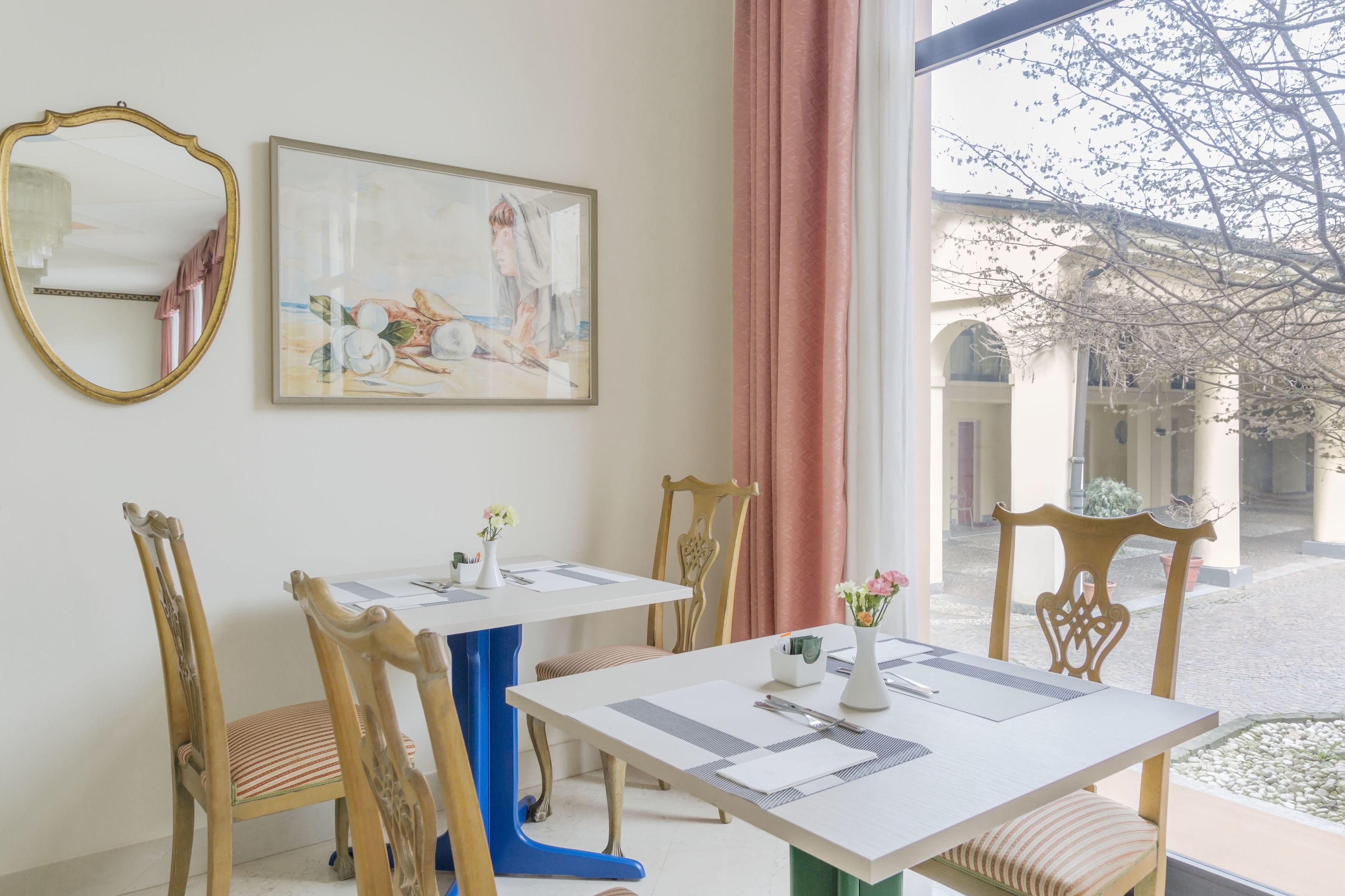 A dining area with a white table, blue base, and wooden chairs. There is a painting on the wall and a window with a view of a building.