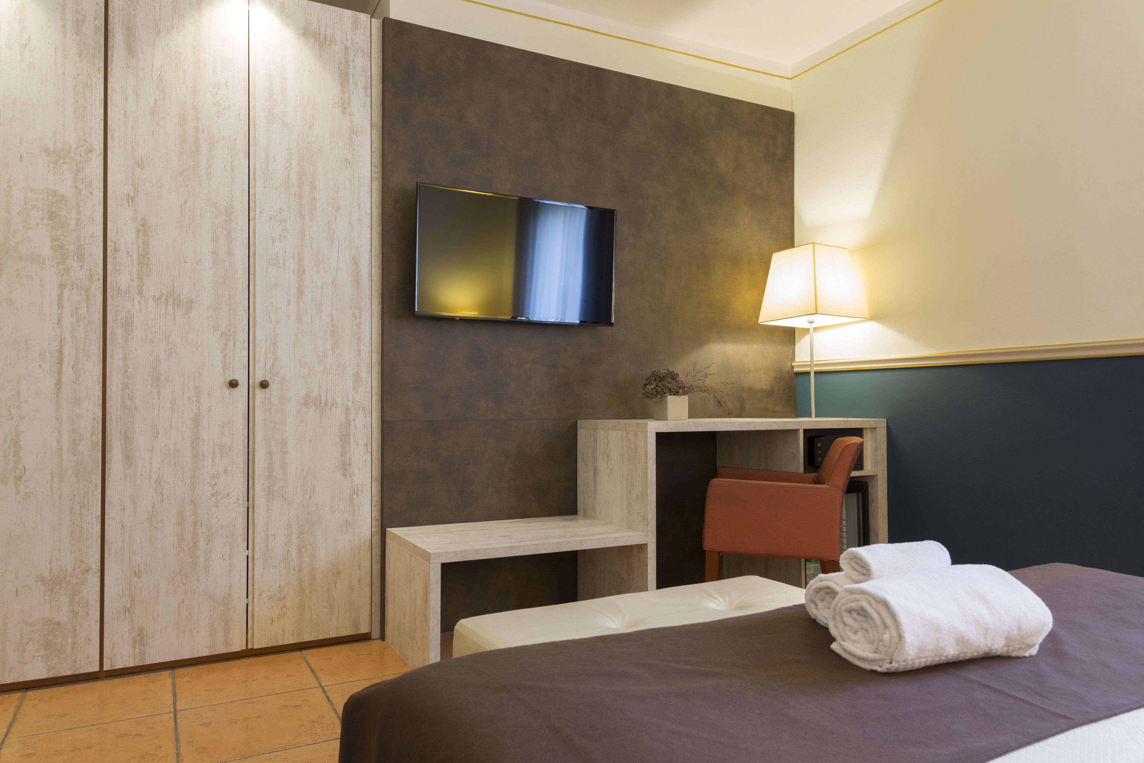 A hotel room with a bed, a TV mounted on the wall, a desk with a chair, and a lamp.