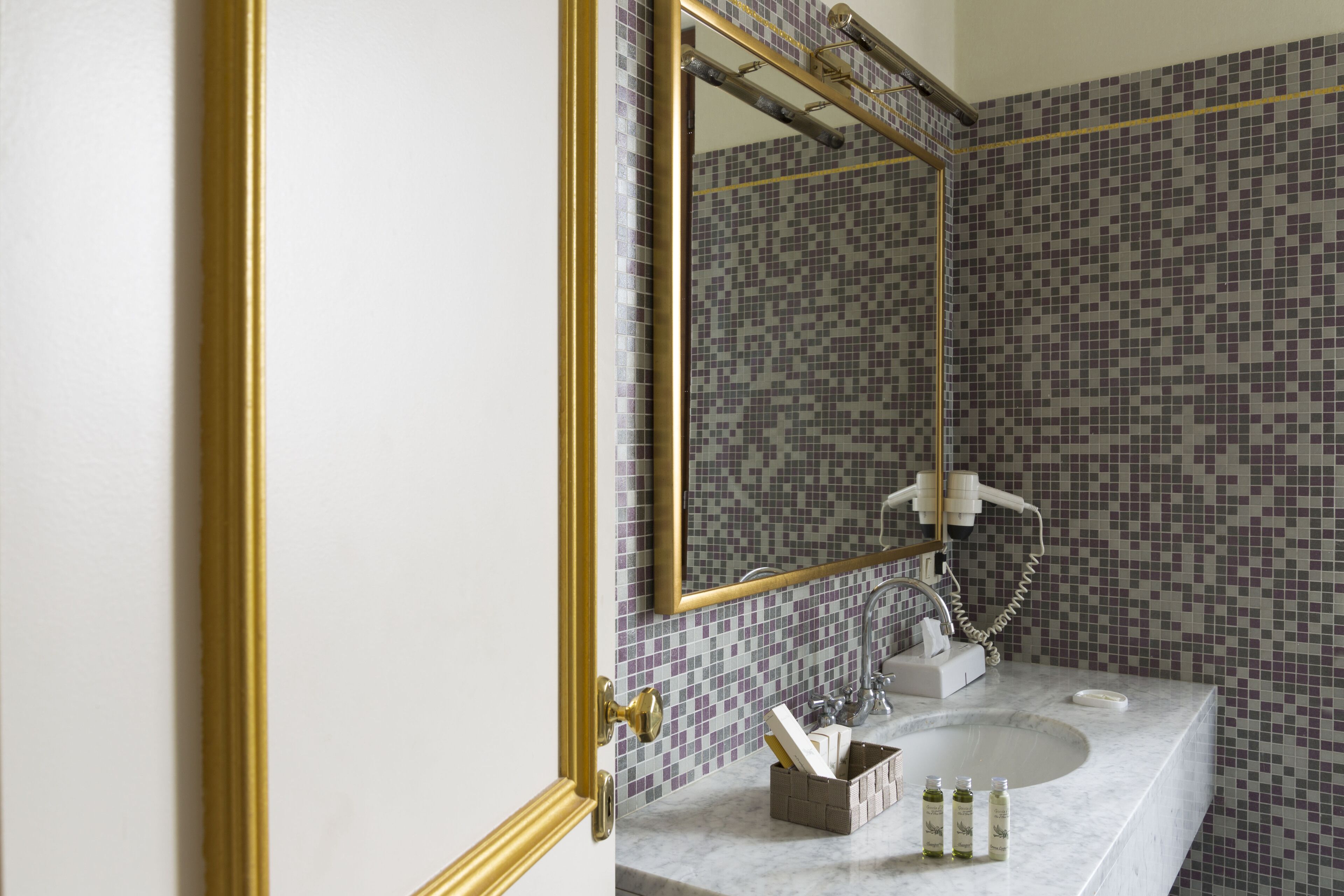 A bathroom with a tiled mosaic wall, a gold-framed mirror, a white sink, and a towel rack.