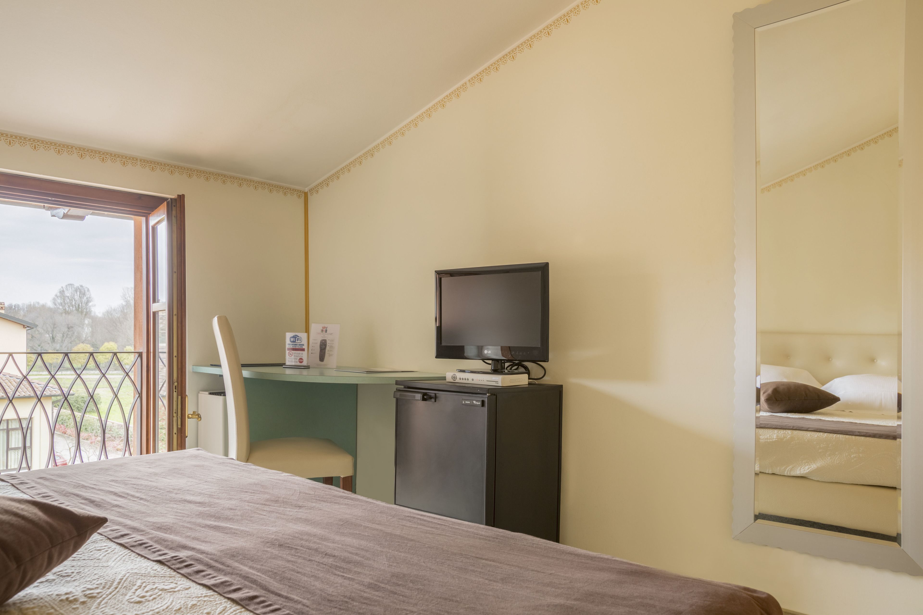 Habitación de hotel con cama, escritorio con computadora y refrigerador pequeño, silla y espejo grande.