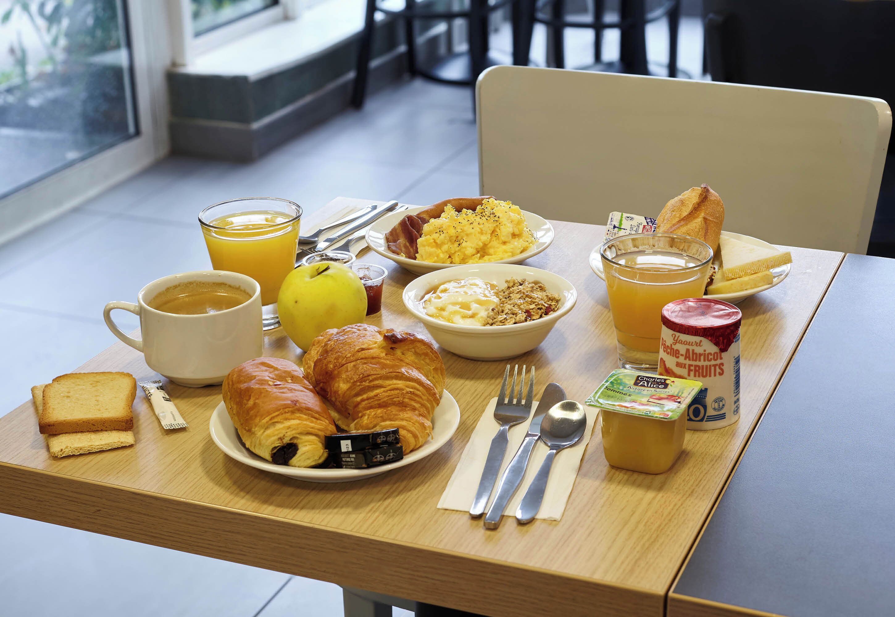 A breakfast table with croissants, toast, a bowl of cereal, a glass of orange juice, a cup of coffee, and a container of yogurt.